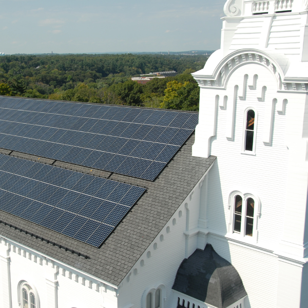 White church building with solar panels on roof and green trees in the background under a cloudy sky. Photo of South Church in Andover, MA.