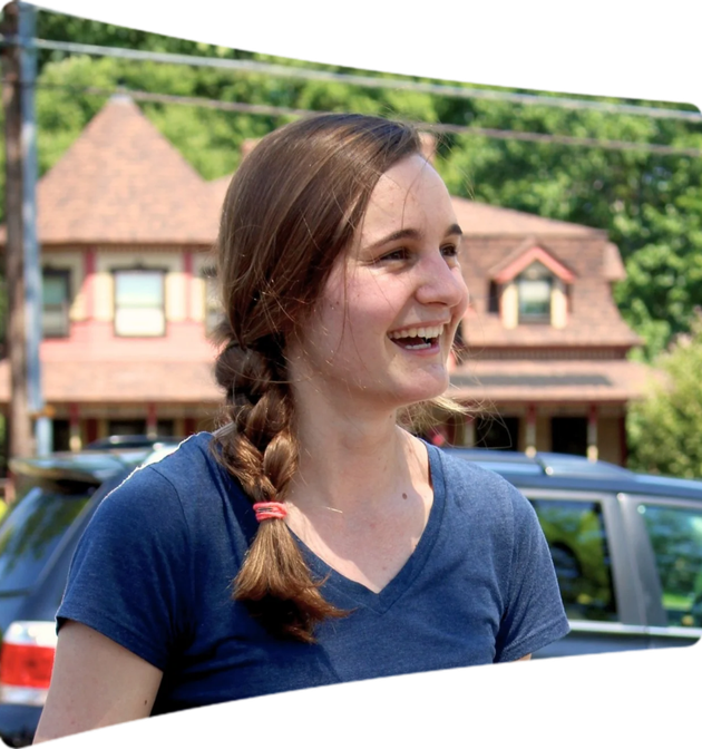 Smiling woman with braided hair wearing a blue shirt outdoors with a house and car in the background.