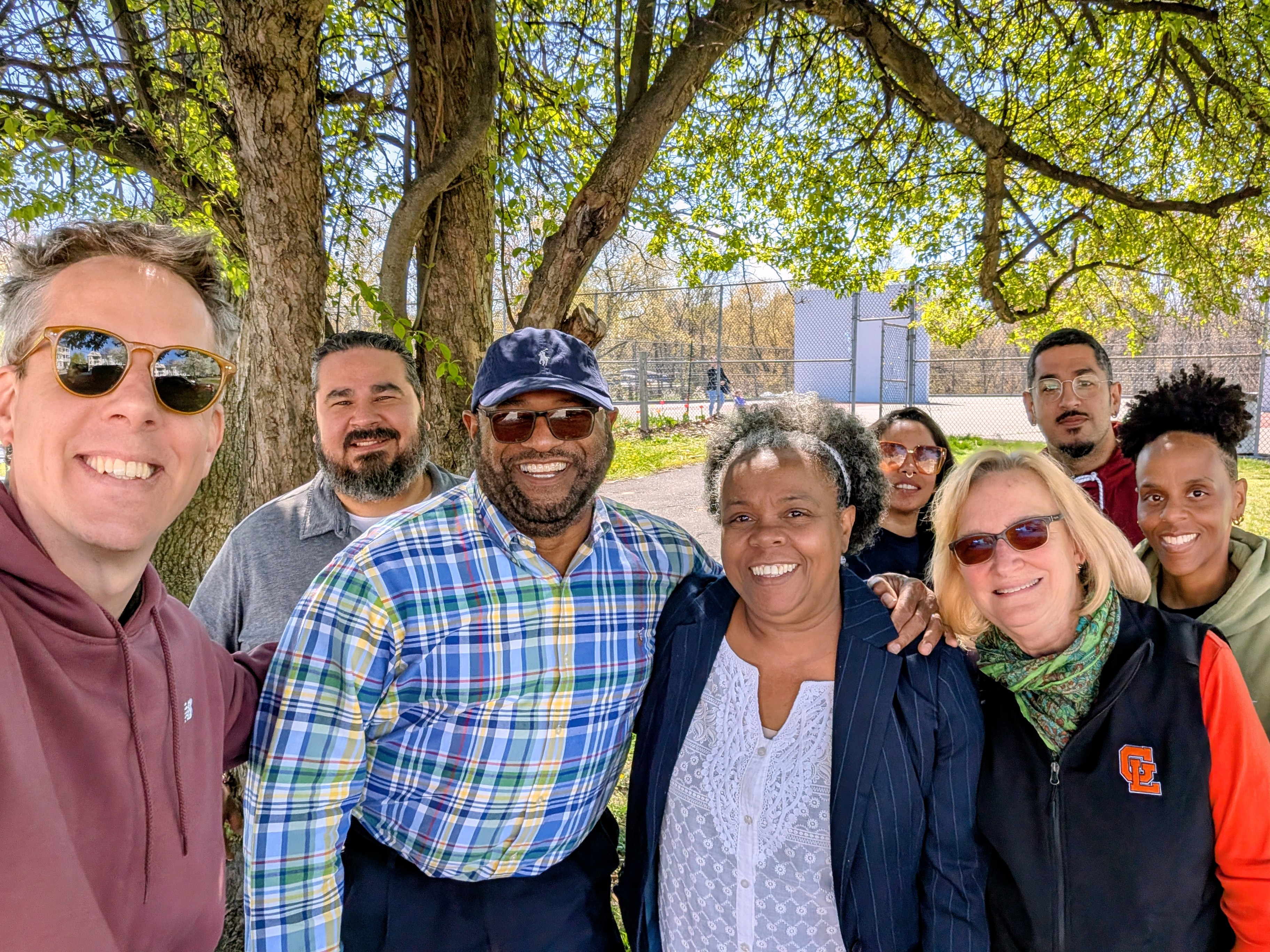 Group of eight diverse adults smiling outdoors under trees on a sunny day.