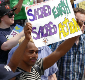Man holding a sign that reads 'Sacred Earth Sacred Action' at a public protest.
