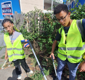 Two Youth Ambassadors in yellow safety vests picking up trash outdoors with grabbers near a fence and bushes.
