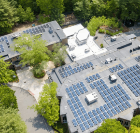 Aerial view of synagogue with solar panels on rooftops surrounded by trees and pathways.