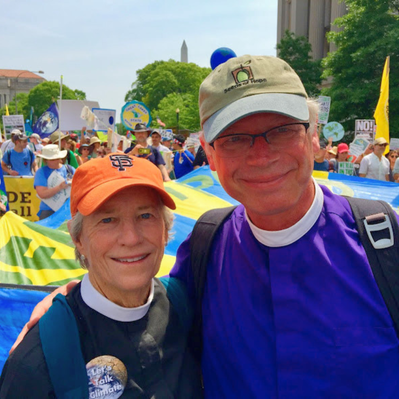 Two people in clergy collars smile at a climate rally with banners and crowds behind them.