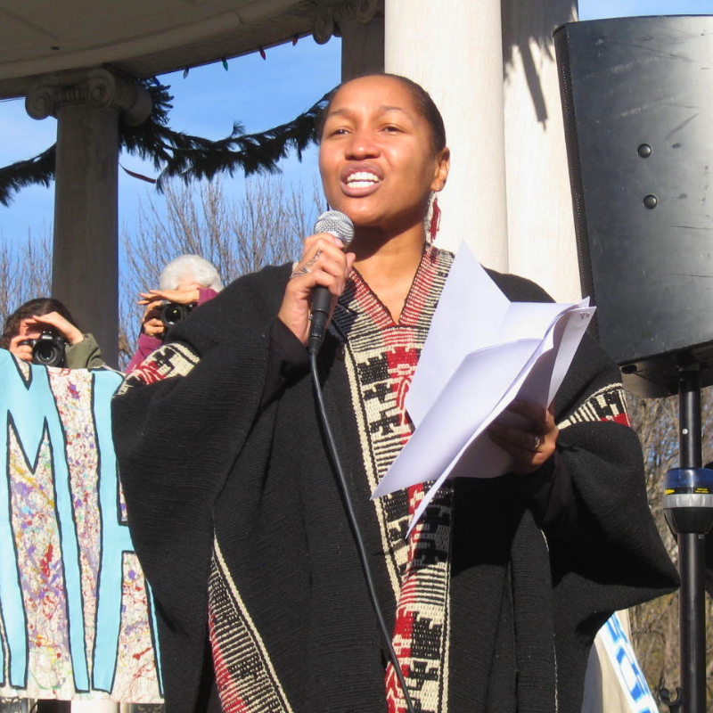 Rev. Mariama White-Hammond
speaking into microphone outdoors, holding papers, with photographers and columns in background.