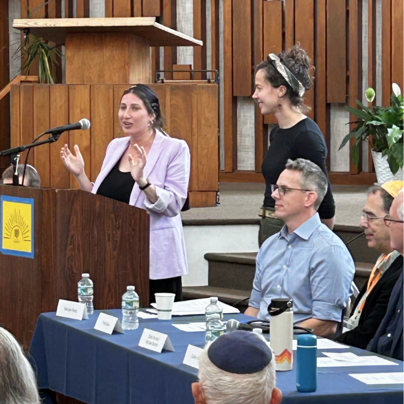 Woman speaking at lectern with seated panelists and audience in a wooden paneled room.