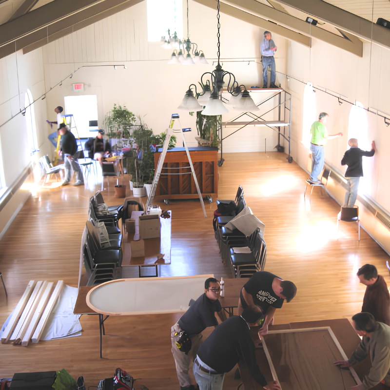 Photo of a MassIPL 'barn raising.' People renovating a spacious sanctuary with wooden floors, assembling wood frames, and working on walls.
