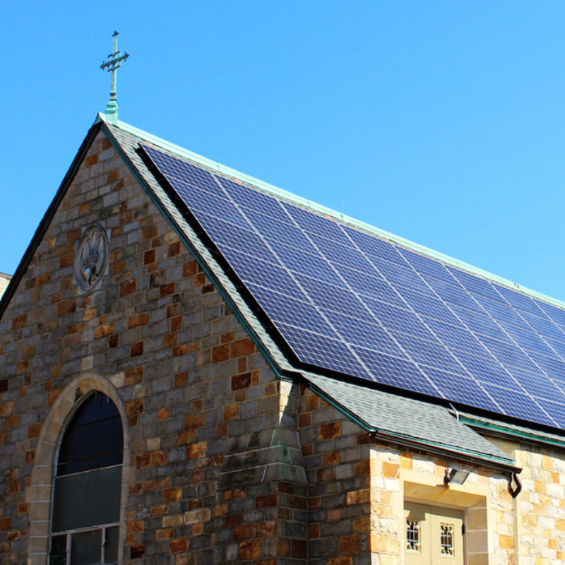 Stone church building with a roof covered in solar panels and a cross at the top against a blue sky.