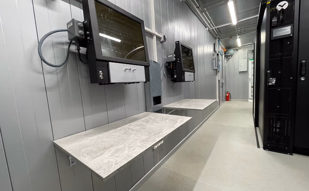 Industrial workspace with two mounted monitors above stainless steel workbenches along a gray paneled wall.