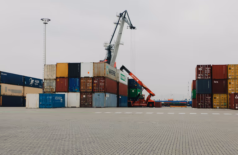 Shipping containers stacked at a port with cranes and machinery under an overcast sky.