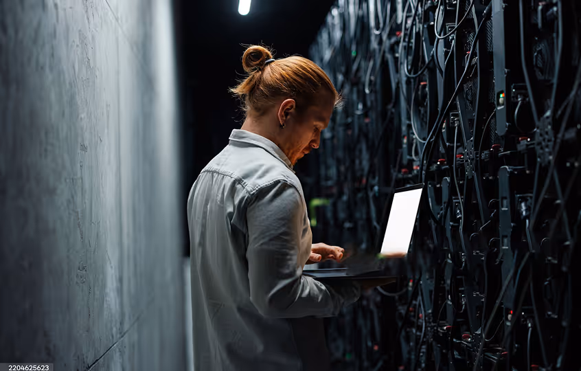 Man with a ponytail standing in a dark server room working on a laptop.