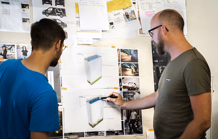 Two men studying and sketching a rectangular design taped on a wall covered with photos and notes.