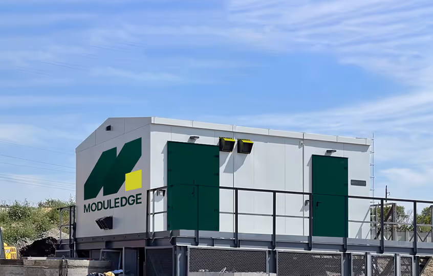 White and green modular building unit with 'MODULEDGE' logo on the side under a clear blue sky.