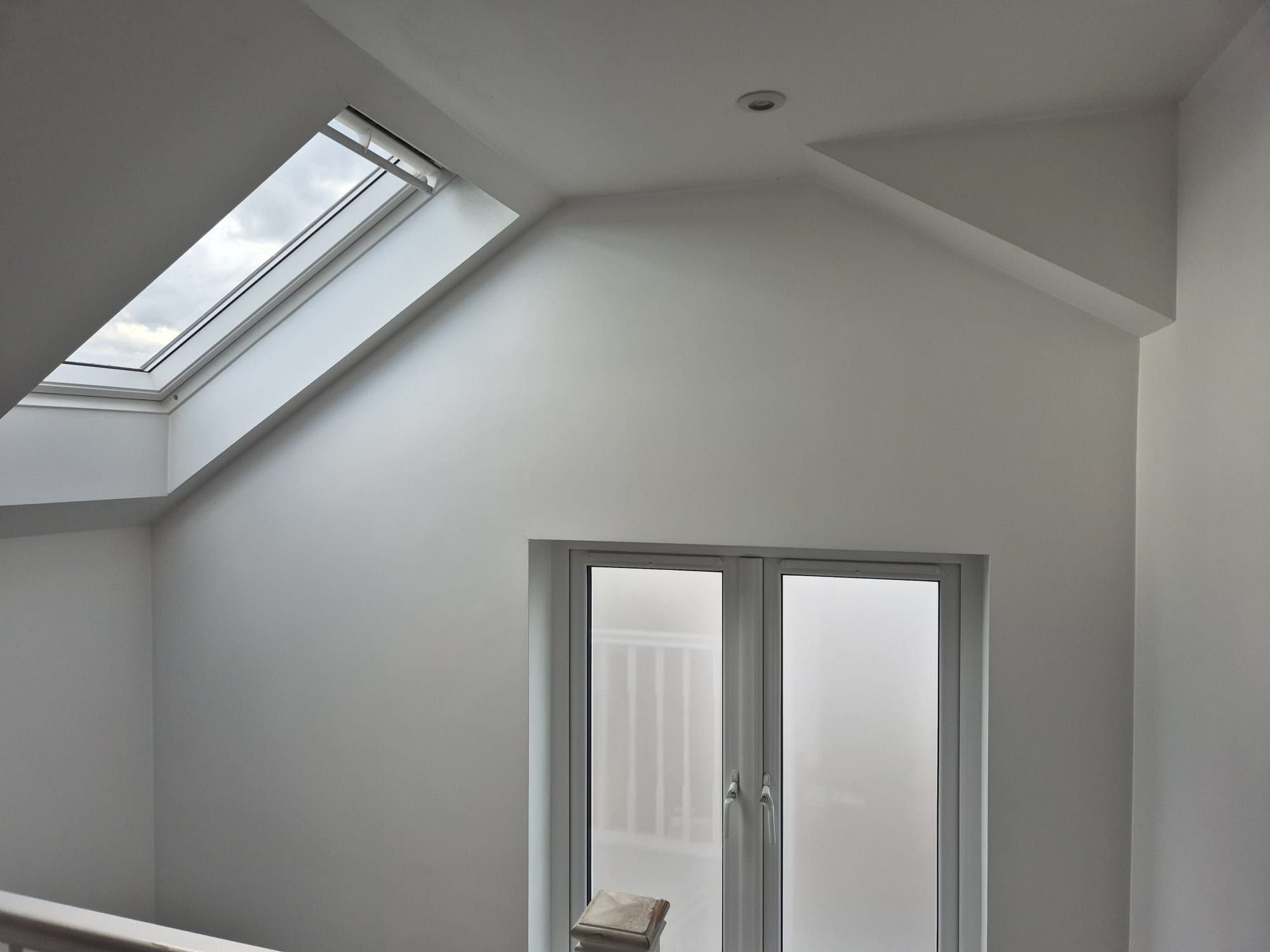 Interior corner of a white room with a slanted ceiling, a skylight window, and a double frosted glass window below.
