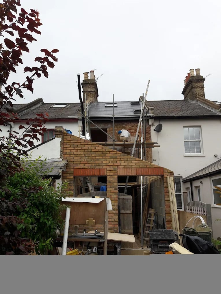 Rear view of a brick house under construction with scaffolding and two workers on the upper level, with construction materials and tools around.