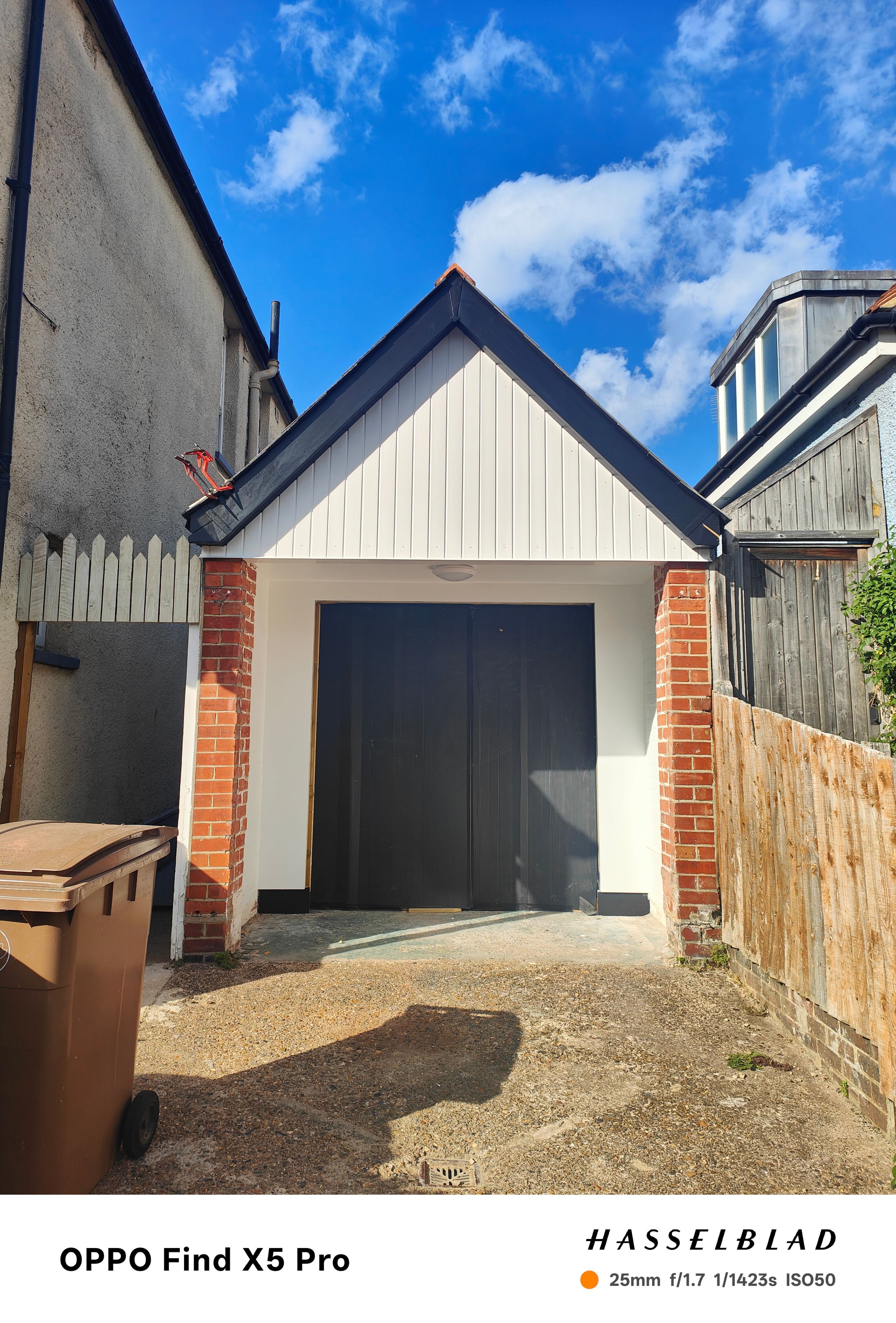 Front view of a small garage with black double doors, red brick pillars, white paneling, and a brown wheelie bin on the left side under a blue sky with clouds.