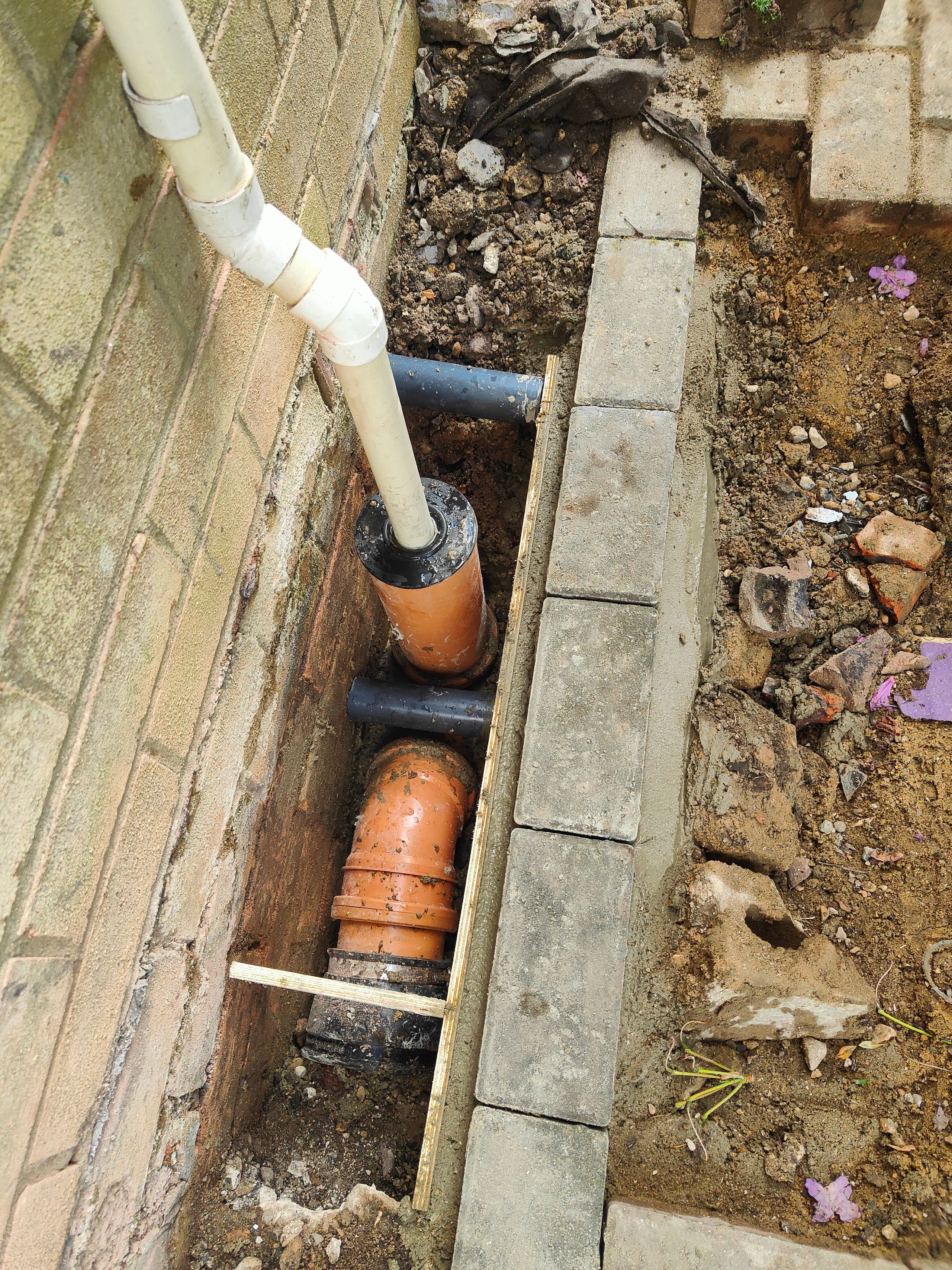 Construction site with exposed drainage pipes next to a brick wall and partially laid stone bricks on dirt.