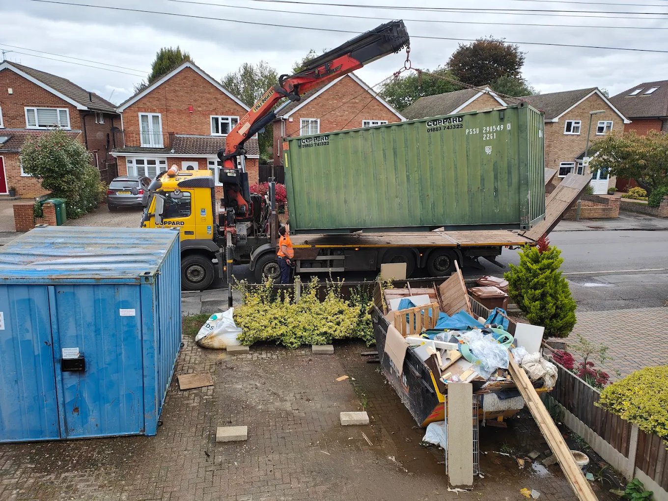 Yellow truck with crane lifting a green shipping container on a residential street with brick houses and a skip filled with debris.