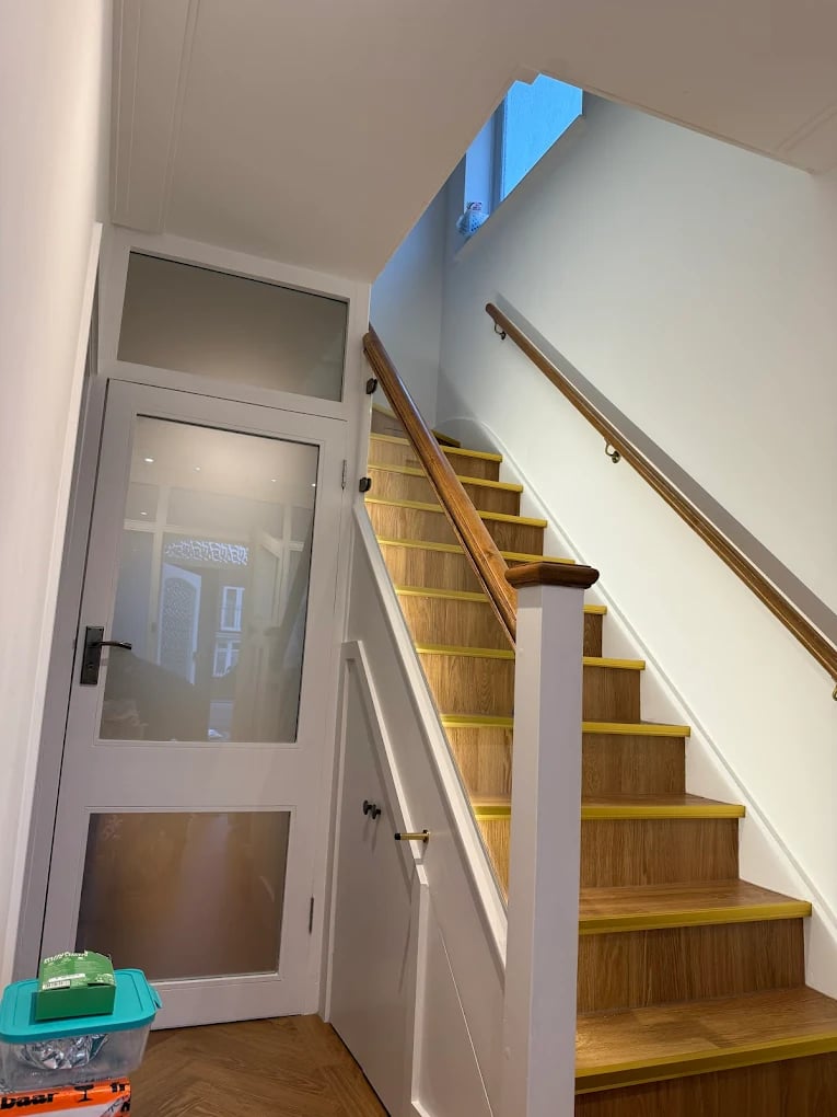 Interior wooden staircase with yellow-edged steps and handrails, next to a white door with frosted glass panels and under-stairs storage.