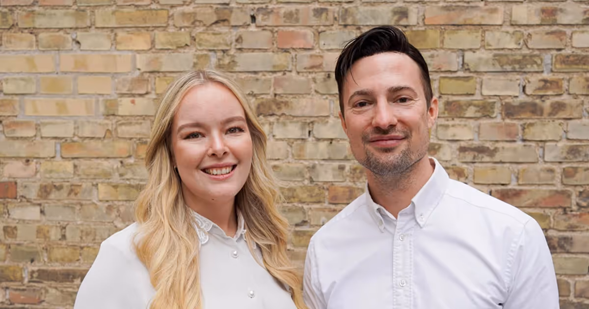 To smilende personer, en kvinde med langt blondt hår og en mand med kort mørkt hår, begge iført hvide skjorter foran en murstensvæg.