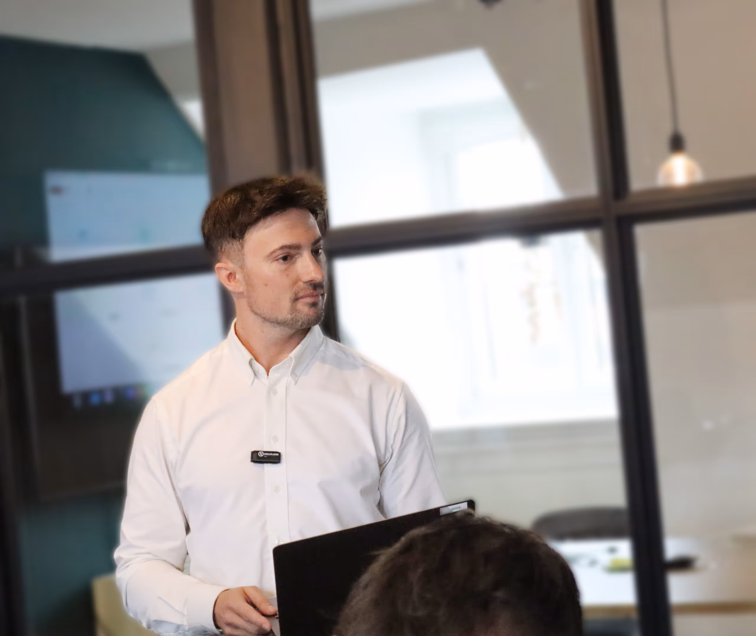 Mand i hvid skjorte holder en bærbar computer og ser til siden i et kontormiljø med glasvægge.