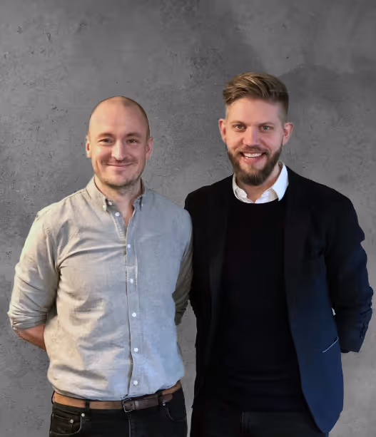 To mænd står smilende foran en grå betonvæg, den ene i lysegrå skjorte og den anden i sort blazer.