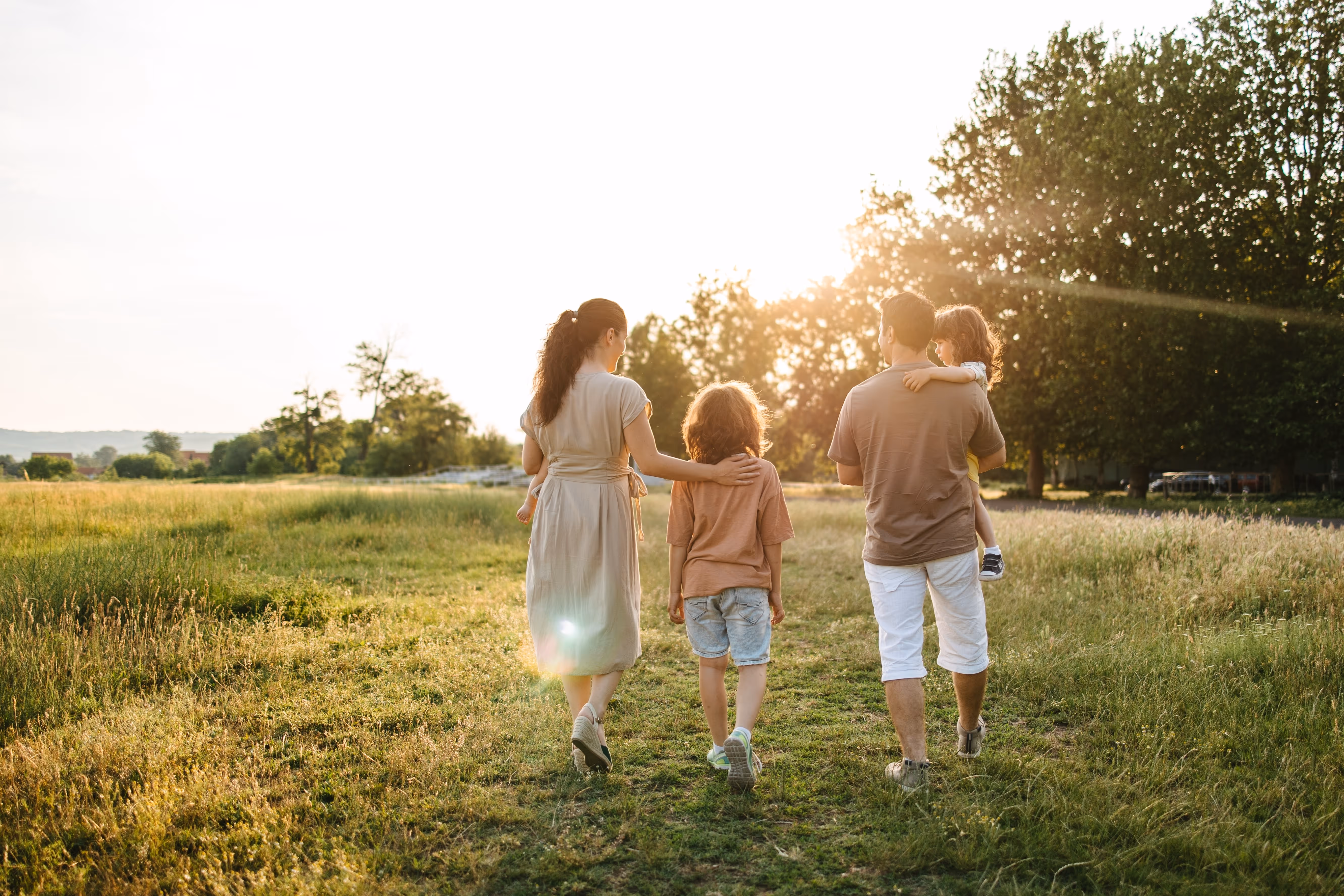 Familie med to voksne og to børn går på en græsvej i solnedgangen omgivet af træer og marker.