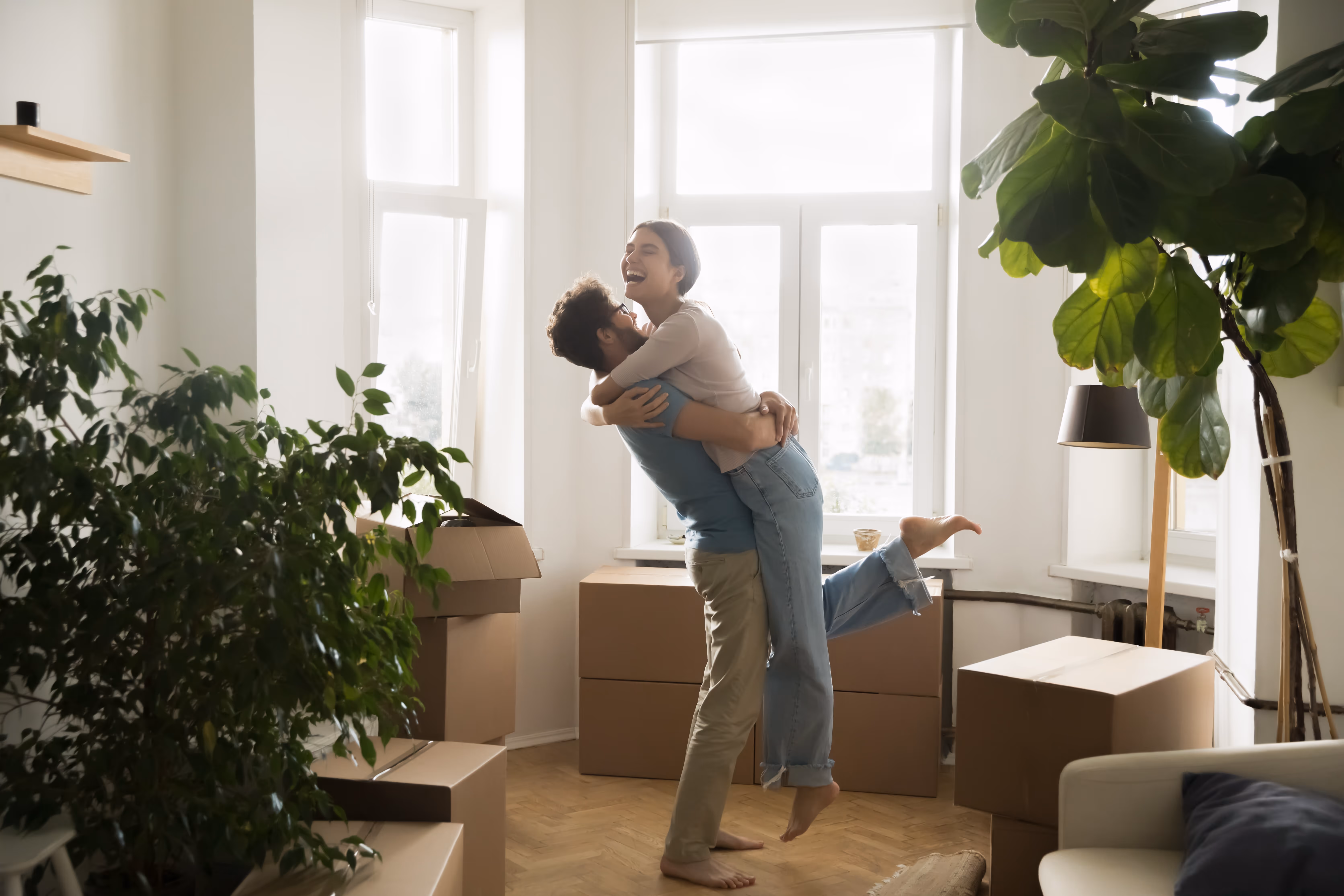 Et par omfavner og smiler lykkeligt i et lyst rum med flyttekasser og planter.