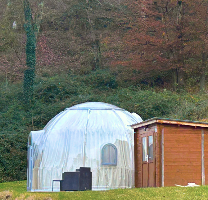 Dôme blanc semi-transparent installé dans un cadre naturel avec un petit bâtiment en bois adjacent et des arbres en arrière-plan.