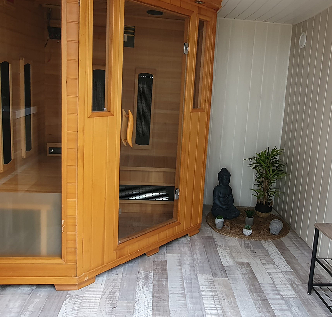 Cabine de sauna en bois avec une porte vitrée dans une pièce au sol en bois clair, près d’un petit coin décoré avec une statue de Bouddha et des plantes.