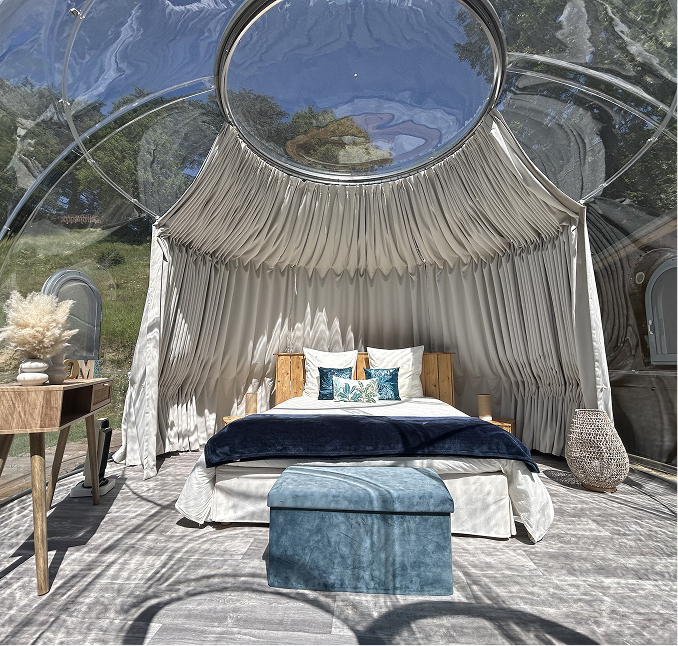 Chambre avec lit double décoré de coussins et couverture bleue dans une tente bulle transparente avec rideaux beige plissés.