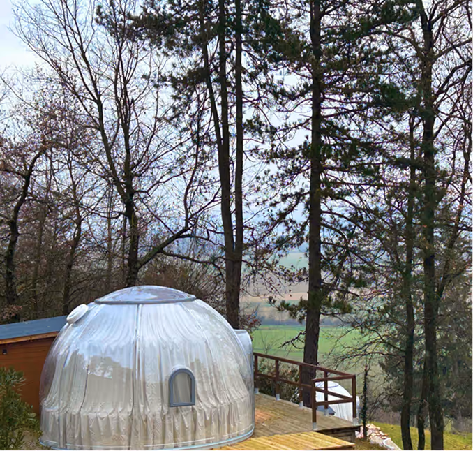 Dôme transparent avec petite fenêtre sur une terrasse en bois entourée d'arbres en hiver.