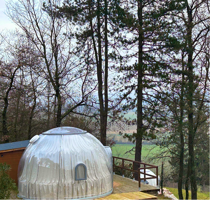 Dôme transparent avec petite fenêtre sur une terrasse en bois entourée d'arbres en hiver.