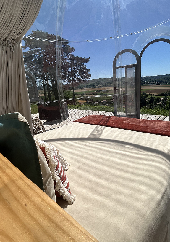 Intérieur d’une bulle transparente avec un lit rond, coussins décoratifs, rideaux beiges, et vue sur la campagne ensoleillée.