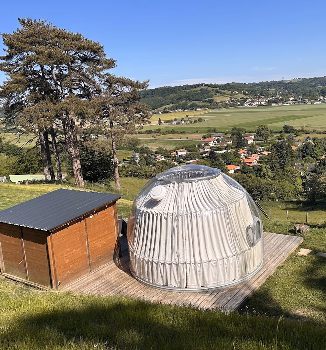 Une bulle d'hébergement transparente avec rideaux blancs sur une terrasse en bois à côté d'un petit abri en bois, entourée d'herbes et d'arbres avec vue sur une campagne vallonnée et un village.