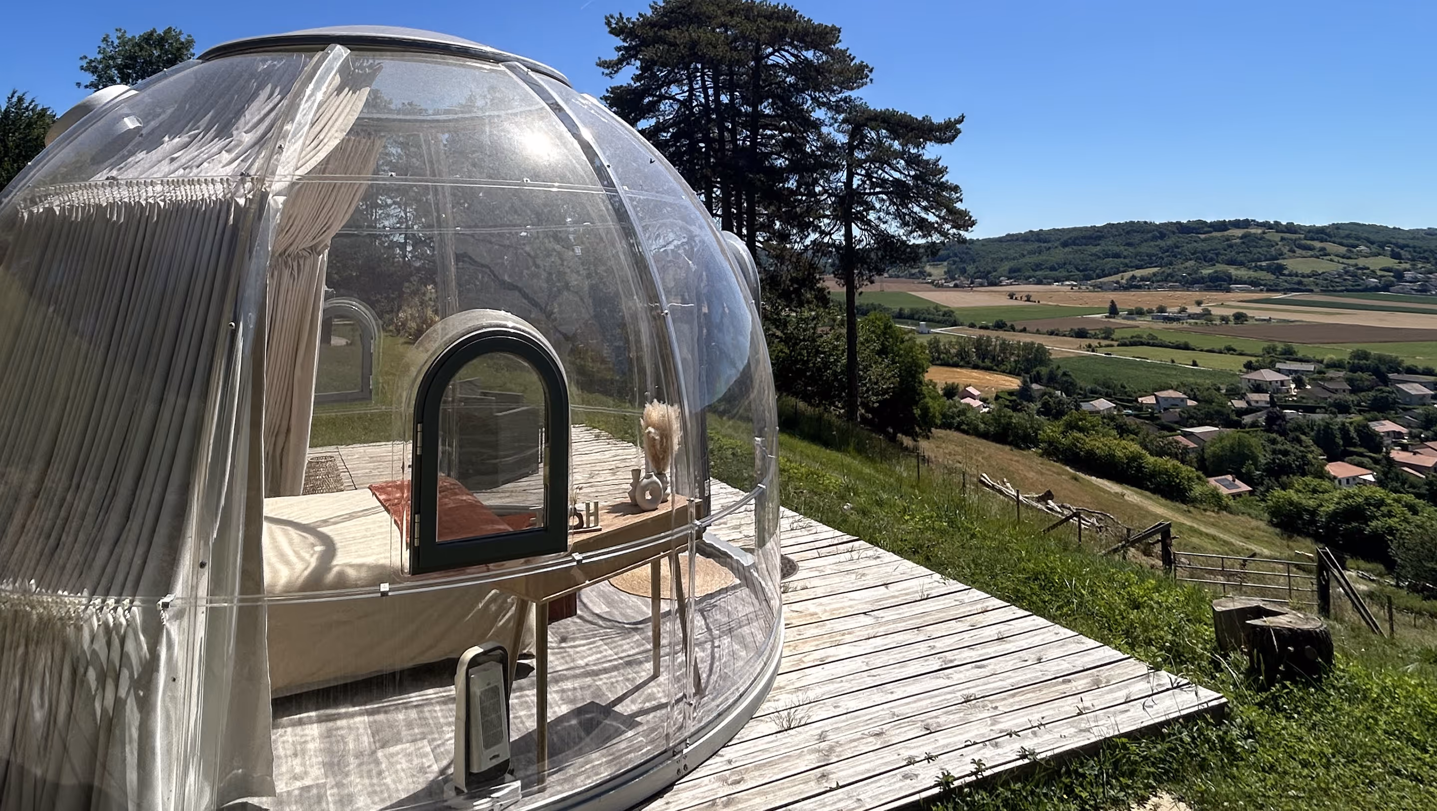Bulle transparente avec un lit et une table à l'intérieur, située sur une terrasse en bois avec vue sur une vallée verte et des collines sous un ciel bleu.