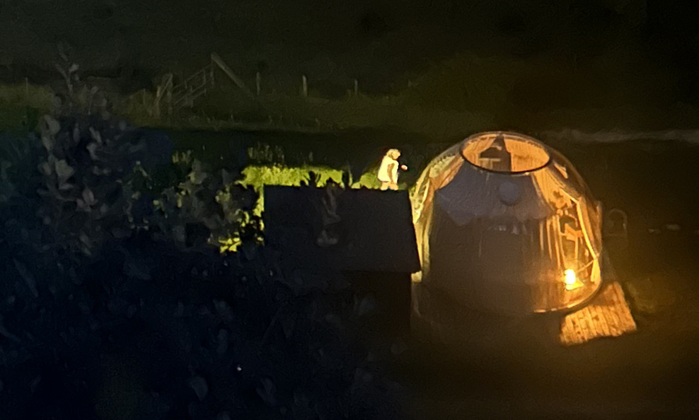 Une tente éclairée la nuit avec une personne debout à proximité sur un pavé, dans un environnement extérieur sombre.