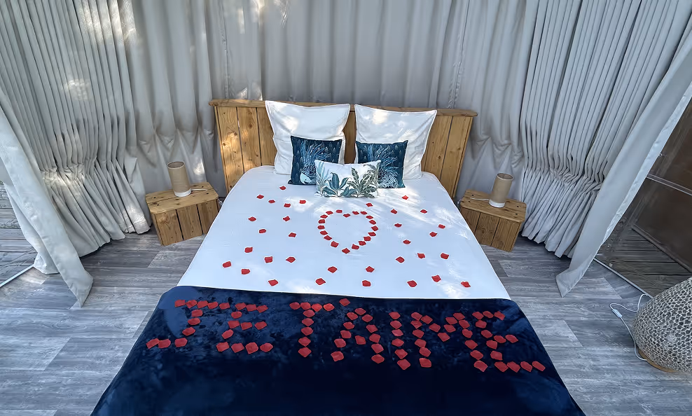 Lit double avec tête de lit en bois, drap blanc décoré de pétales rouges formant un cœur et le message 'JE T'AIME' sur une couverture bleue.