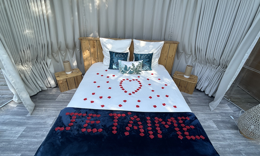 Lit double avec tête de lit en bois, drap blanc décoré de pétales rouges formant un cœur et le message 'JE T'AIME' sur une couverture bleue.
