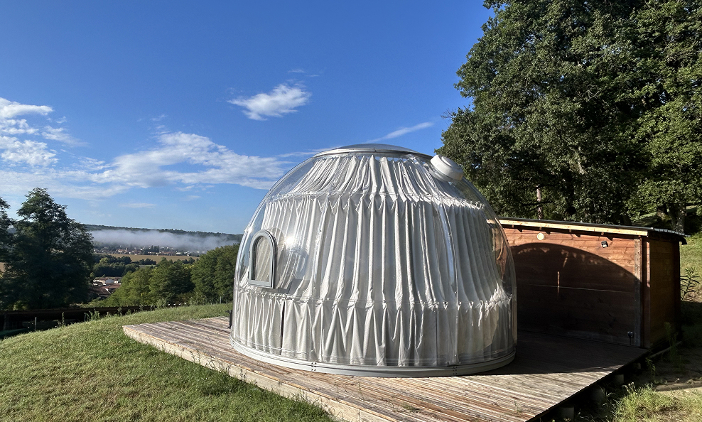 Habitation en forme de dôme transparent avec rideaux blancs, installée sur une terrasse en bois dans une zone verdoyante sous un ciel bleu.