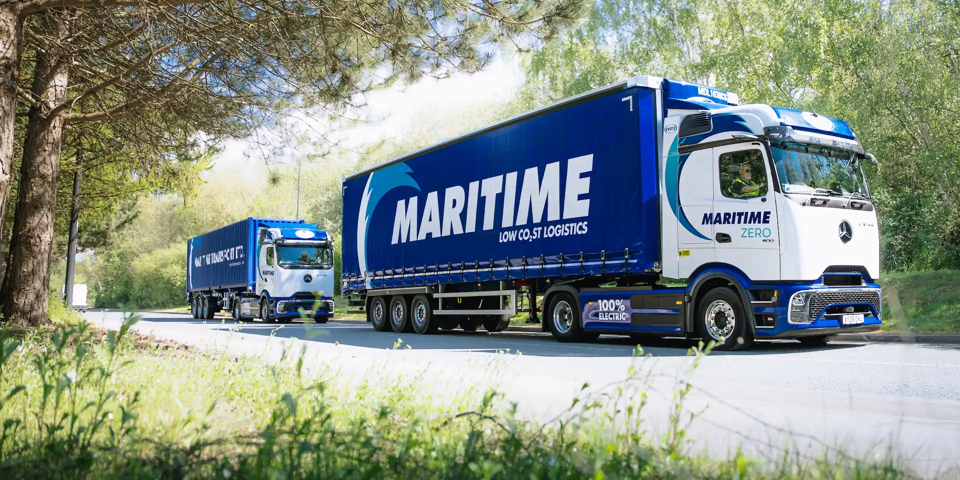 Two Maritime Transport eHGVs driving on a road surrounded by grass and trees.
