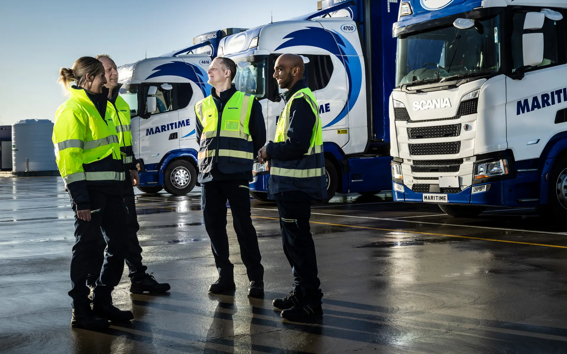 Four drivers in high-visibility vests talk in front of vehicle fleet.