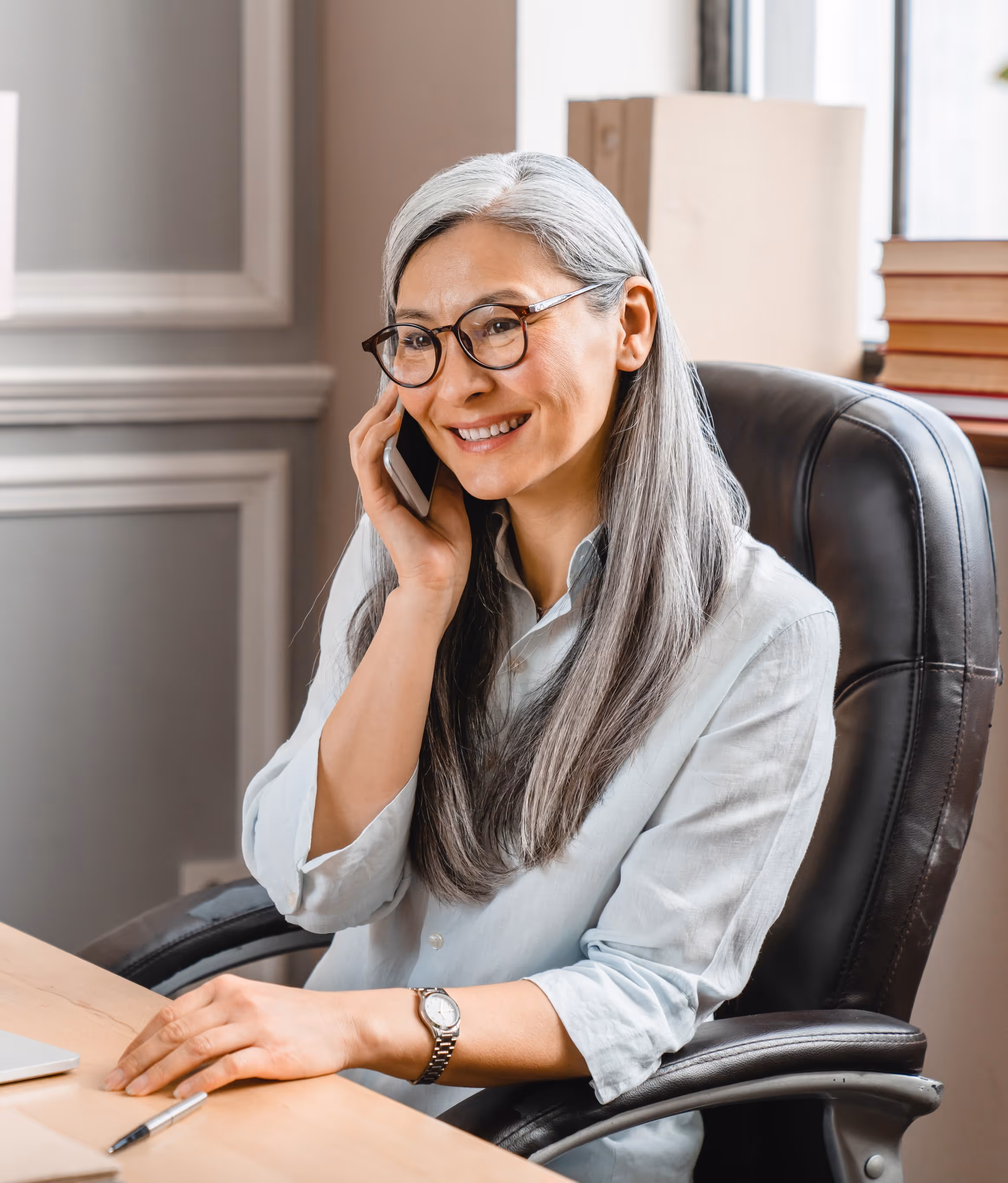 Smiling woman with gray hair and glasses talking on a phone while sitting at an office desk.