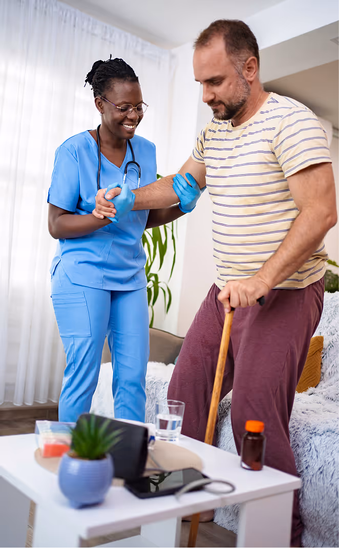 Healthcare worker in blue scrubs assisting an elderly man with a cane as he stands up in a living room.