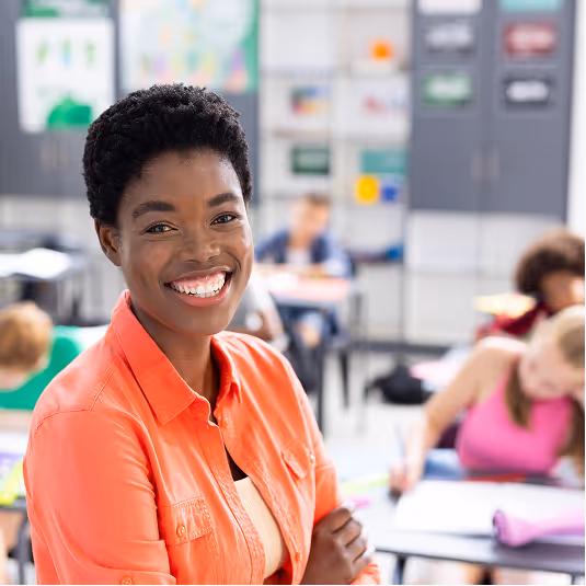 Smiling young black woman in an orange shirt standing in a classroom with students studying in the background.