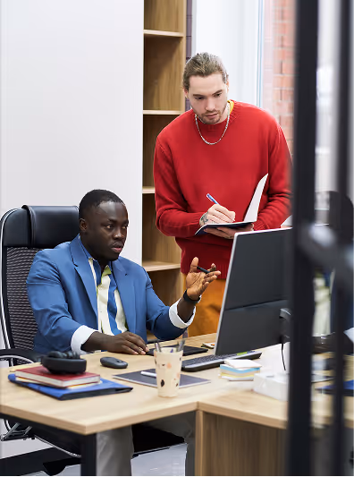 Two professionals collaborating at a desk with a computer; one is seated in a blue suit gesturing towards the screen, while the other in a red sweater takes notes.