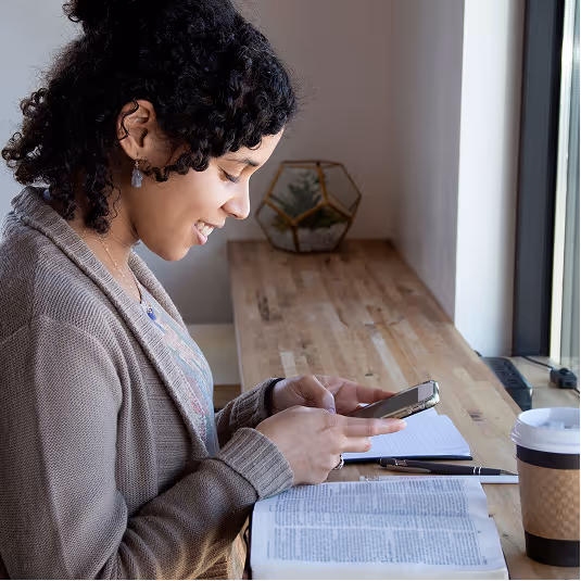 Woman with curly hair sitting at a wooden desk by a window, smiling while using her smartphone with an open book, notebook, pen, and coffee cup in front of her.