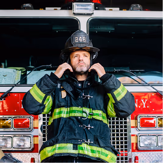 Firefighter adjusting collar of protective gear, standing in front of red fire truck with visible lights and grille.