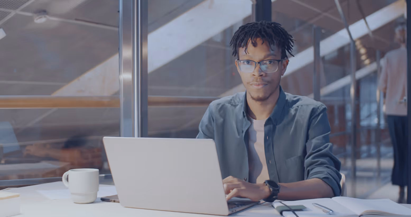 Young man with glasses sitting at a desk typing on a laptop in a modern office with a coffee mug and open notebook nearby.