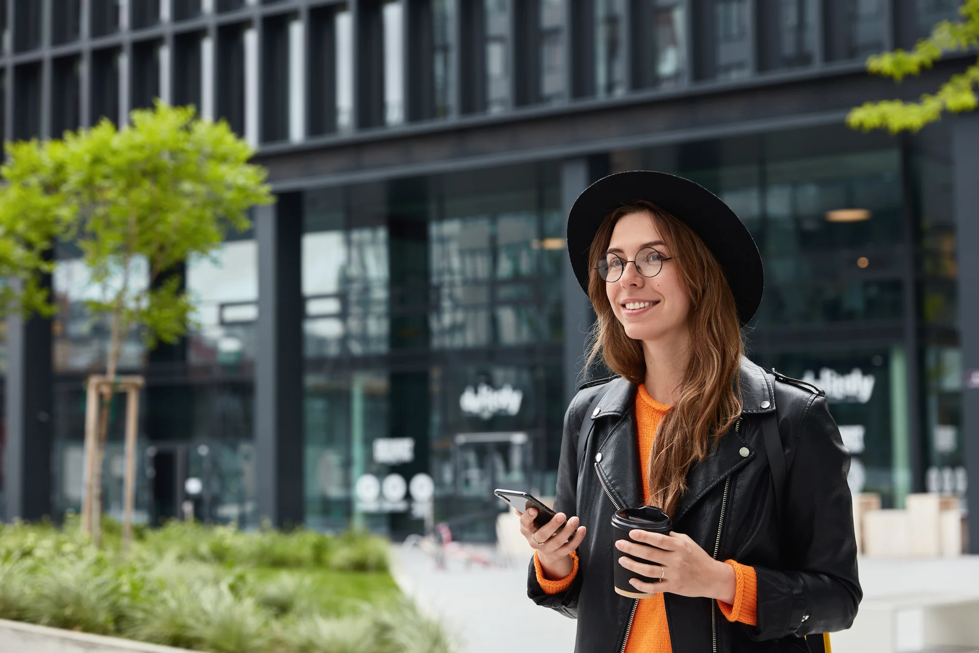 Smiling young woman wearing a black hat and leather jacket holding a smartphone and a takeaway coffee cup outdoors.