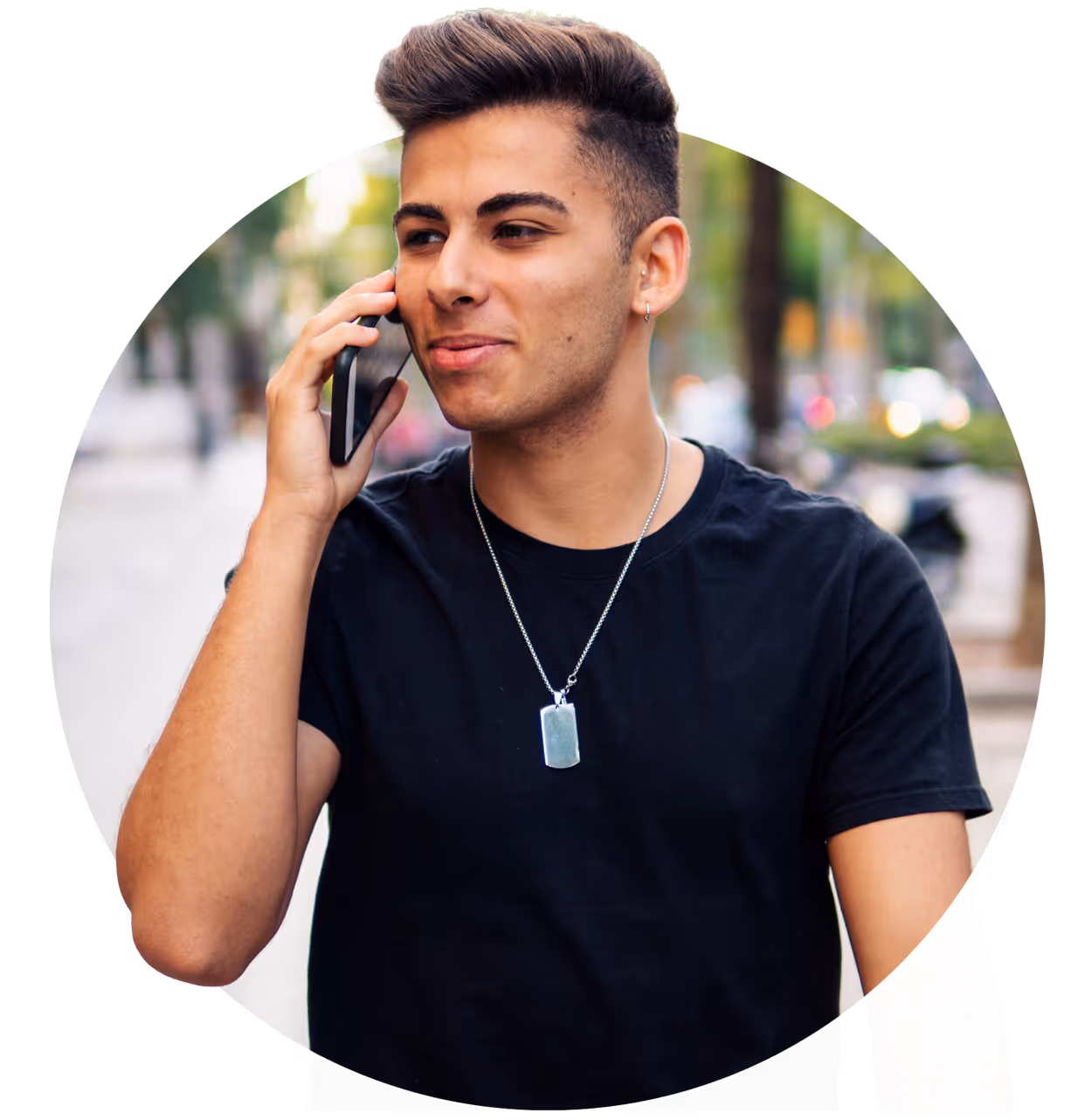 Young man with short hair wearing a black t-shirt and silver pendant talking on a smartphone outdoors.